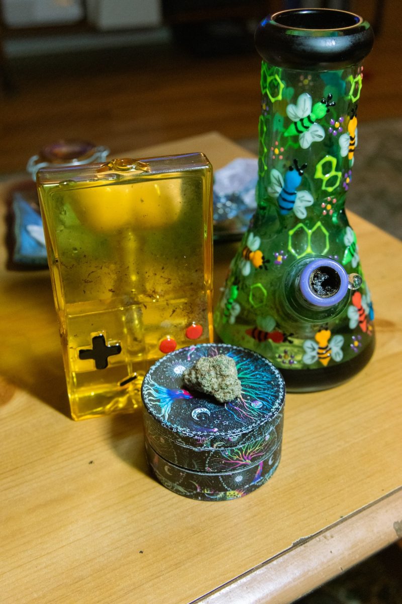 The set up for a magical girls' night with a gameboy water bong and glow-in-the-dark bee/lightning bug water bong and witchy grinder with a nug of Glitter Bomb sitting on top and a tray of crystals and a The Stoner stash jar in the background.