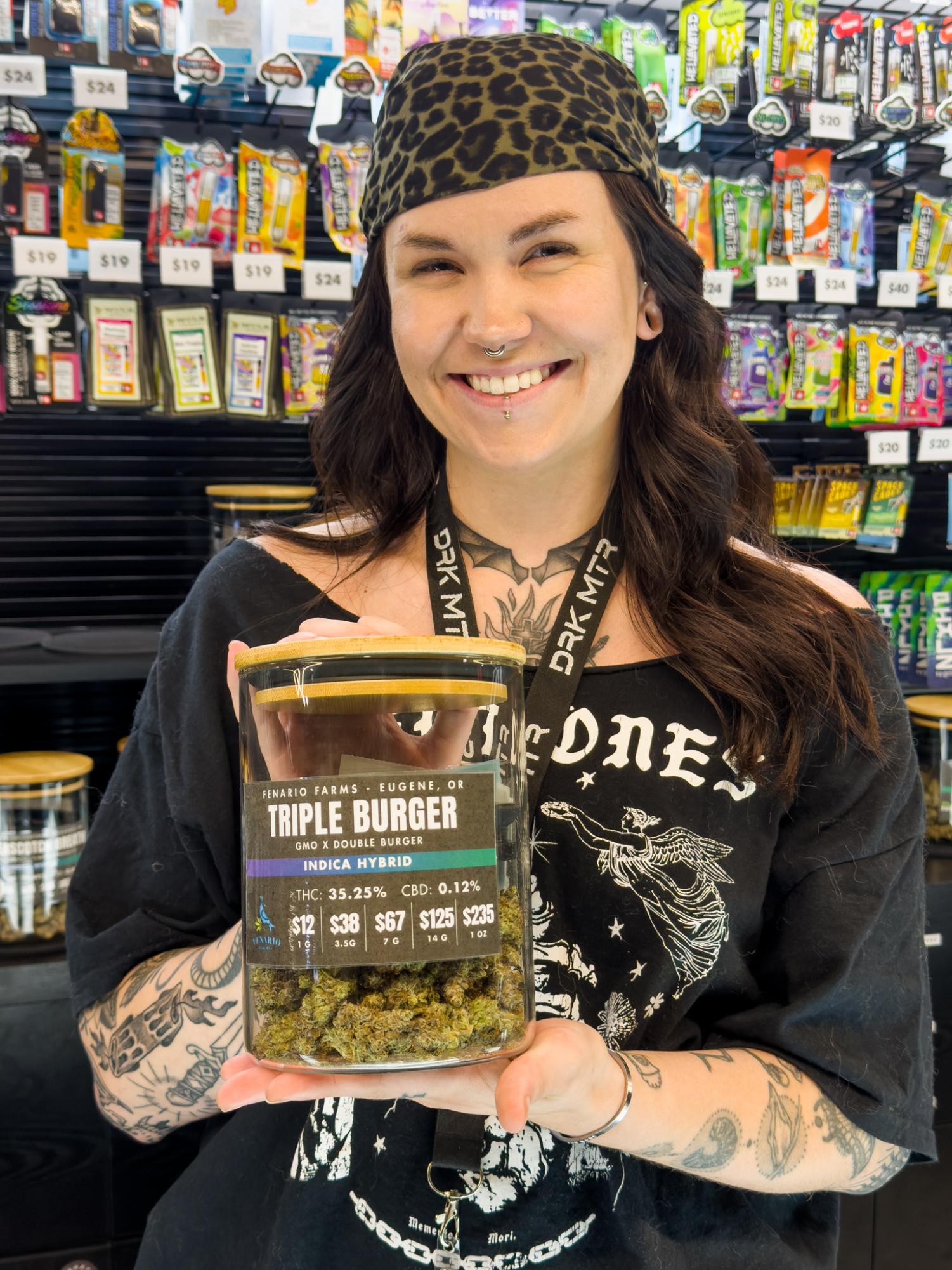 Sage is standing behind the counter at Dark Matter holding a container of Triple Burger flower and smiling at the camera.